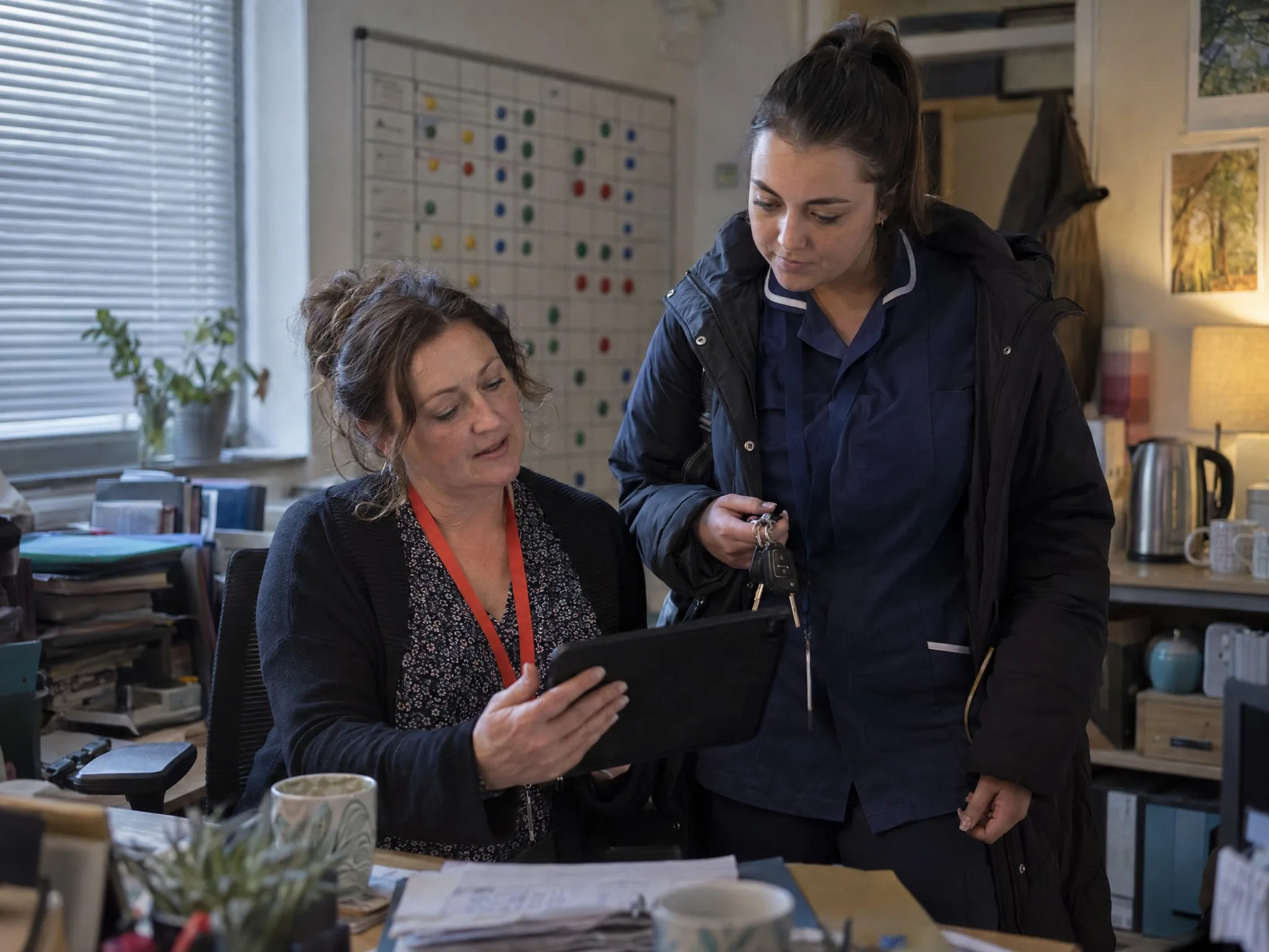 Early morning in a care office: a coordinator shows a carer, coat on and keys in hand, the day's first visit on a tablet.
